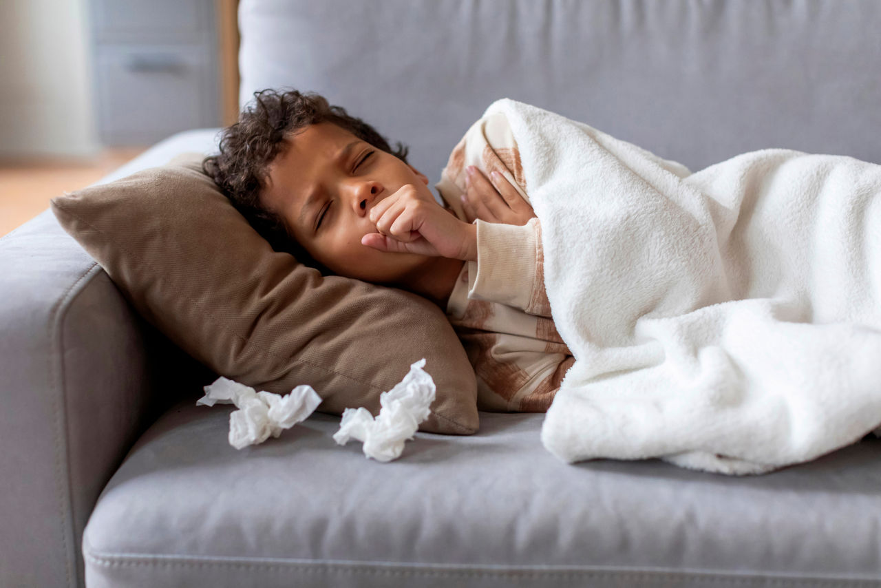 Sick black boy feeling unwell at home, coughing into fist while lying on couch under white blanket with tissues nearby, ill male kid resting during the day in quiet living room, closeup